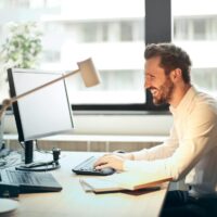 man in white dress shirt sitting on black rolling chair while facing black computer set and smiling