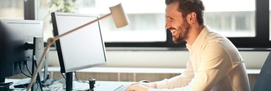 man in white dress shirt sitting on black rolling chair while facing black computer set and smiling
